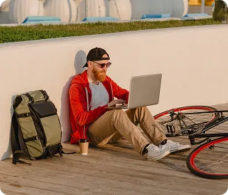 Man in red hoodie sitting on ground using laptop beside a bicycle and backpack outdoors.